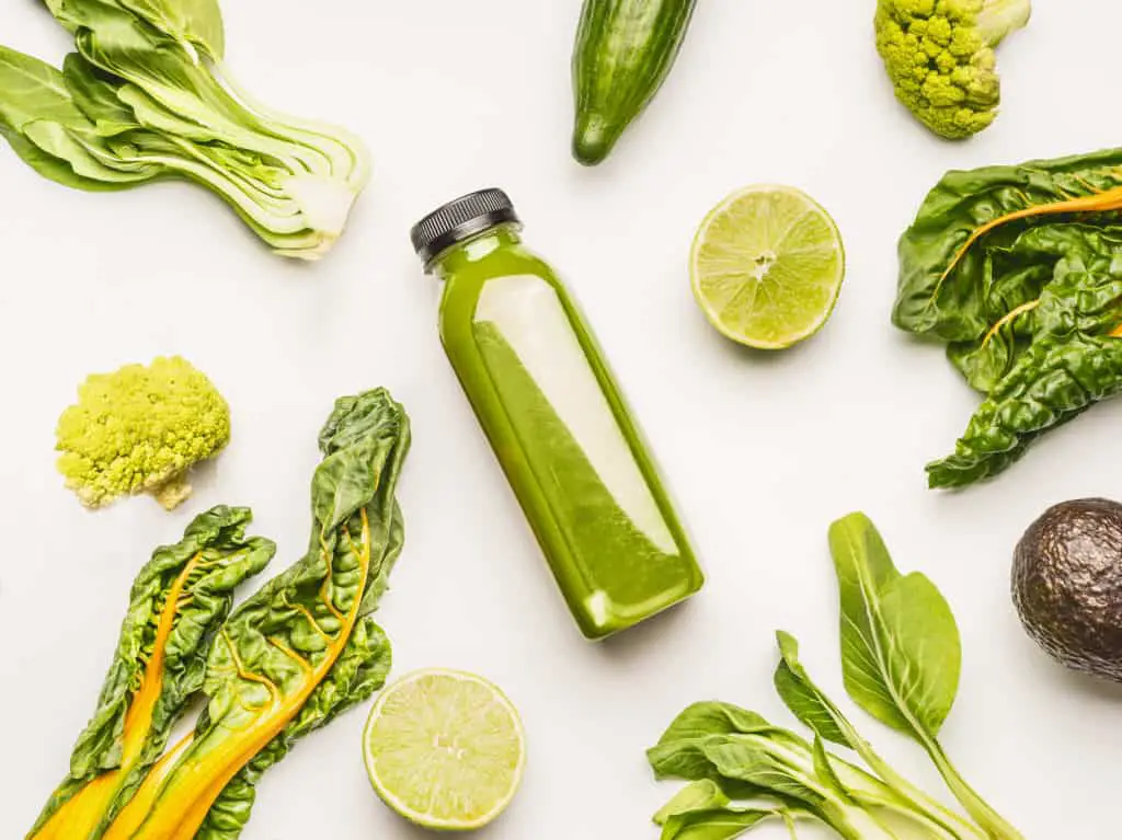 Plastic bottle filled with green juice lays on its side on a white table surrounded by leafy green vegetables