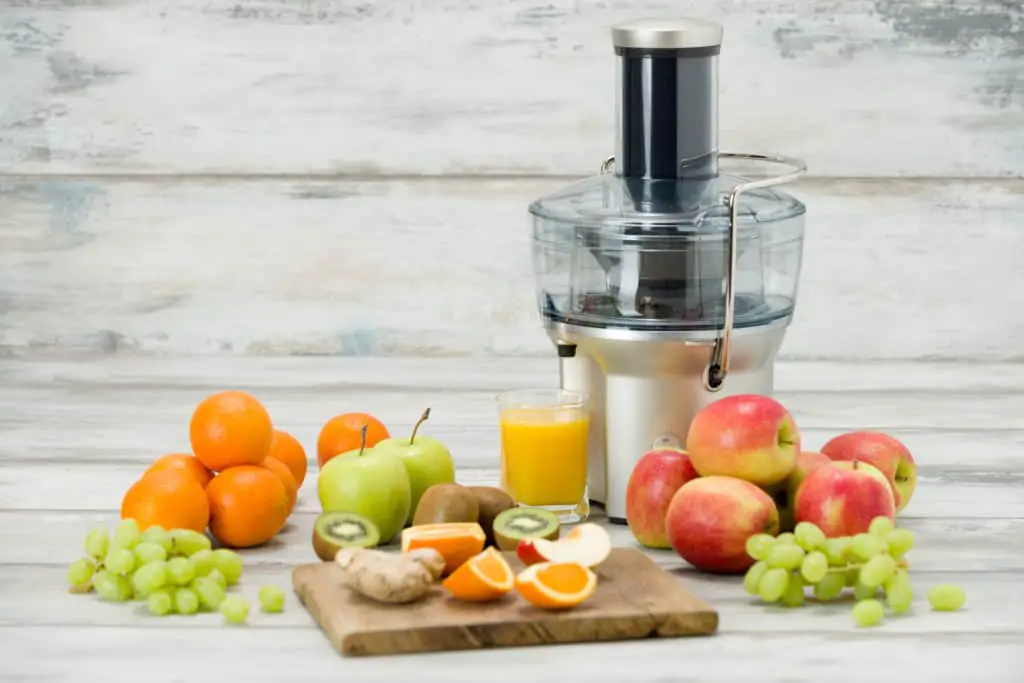 Centrifugal juicer on a wood table surrounded by fruit and a glass of freshly made juice