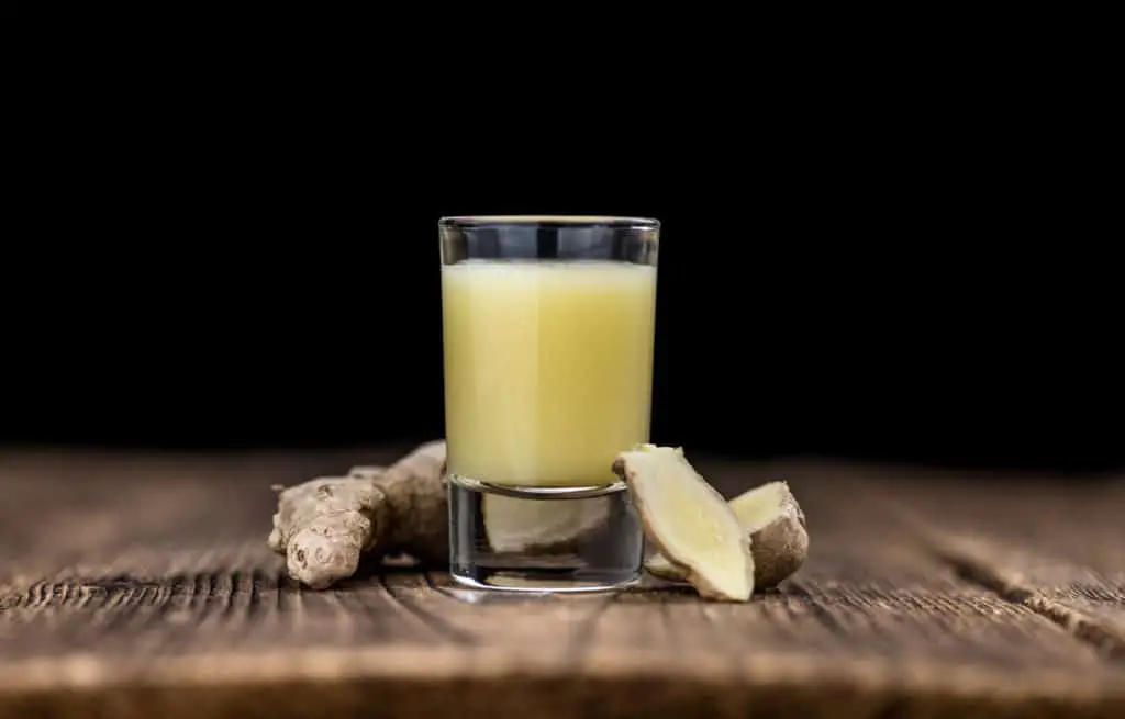 Ginger juice in a clear shot glass surrounded by ginger on a wooden table