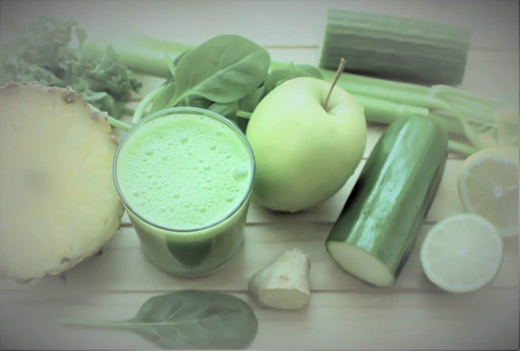 Cucumber, Lemon, and Ginger Juice in a glass cup, surrounded by lemons, cucumbers, apples, celery, and pineapple on a wooden table