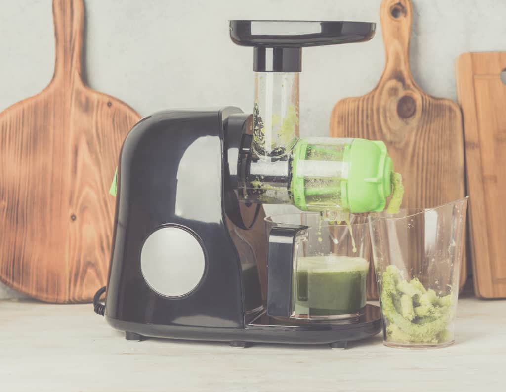 Black horizontal masticating juicer making green juice on top of a white table with wooden cutting boards in the background